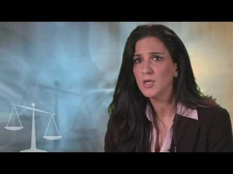 A woman with long dark hair speaks, dressed in a business suit. A faint image of scales is in the lower left corner.