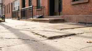 cracked and uneven sidewalk slab near brick building entrance with iron railing